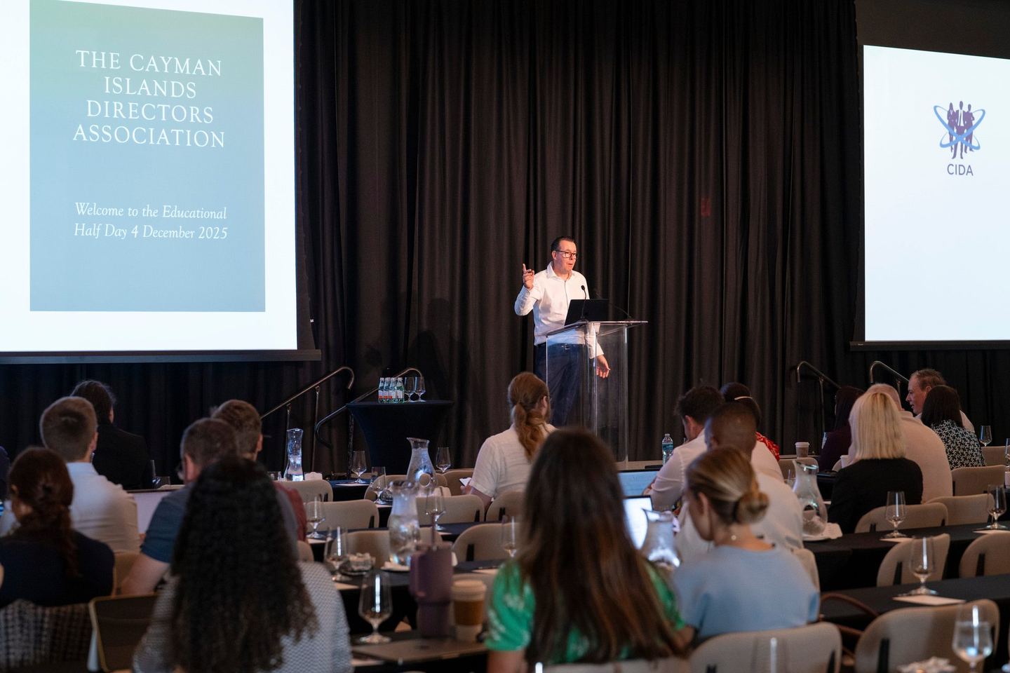 Wide view of the conference room at Hotel Indigo, a speaker at the lectern beside a screen reading “The Cayman Islands Directors Association – Welcome to the Educational Half Day”