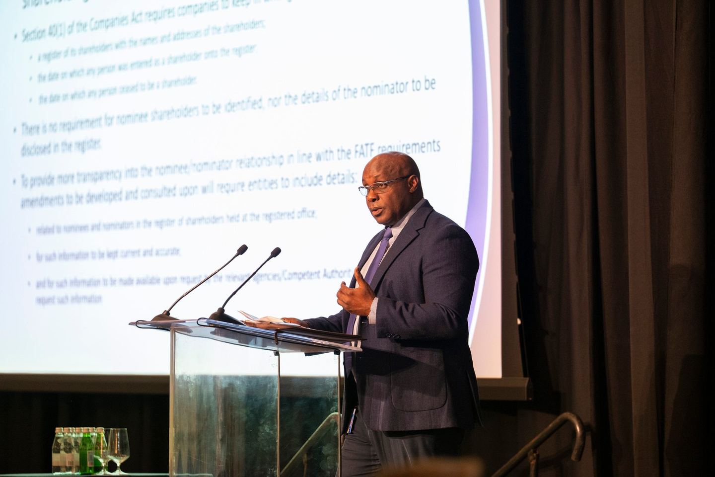 A speaker at a glass lectern in front of a projected slide on the Companies Act
