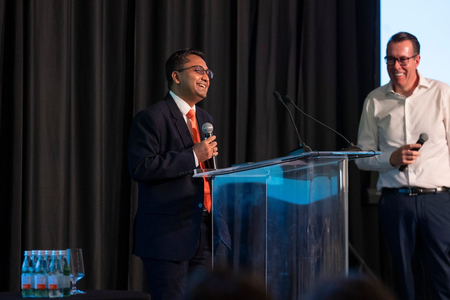 Two presenters on stage, one at the lectern and one standing with a microphone